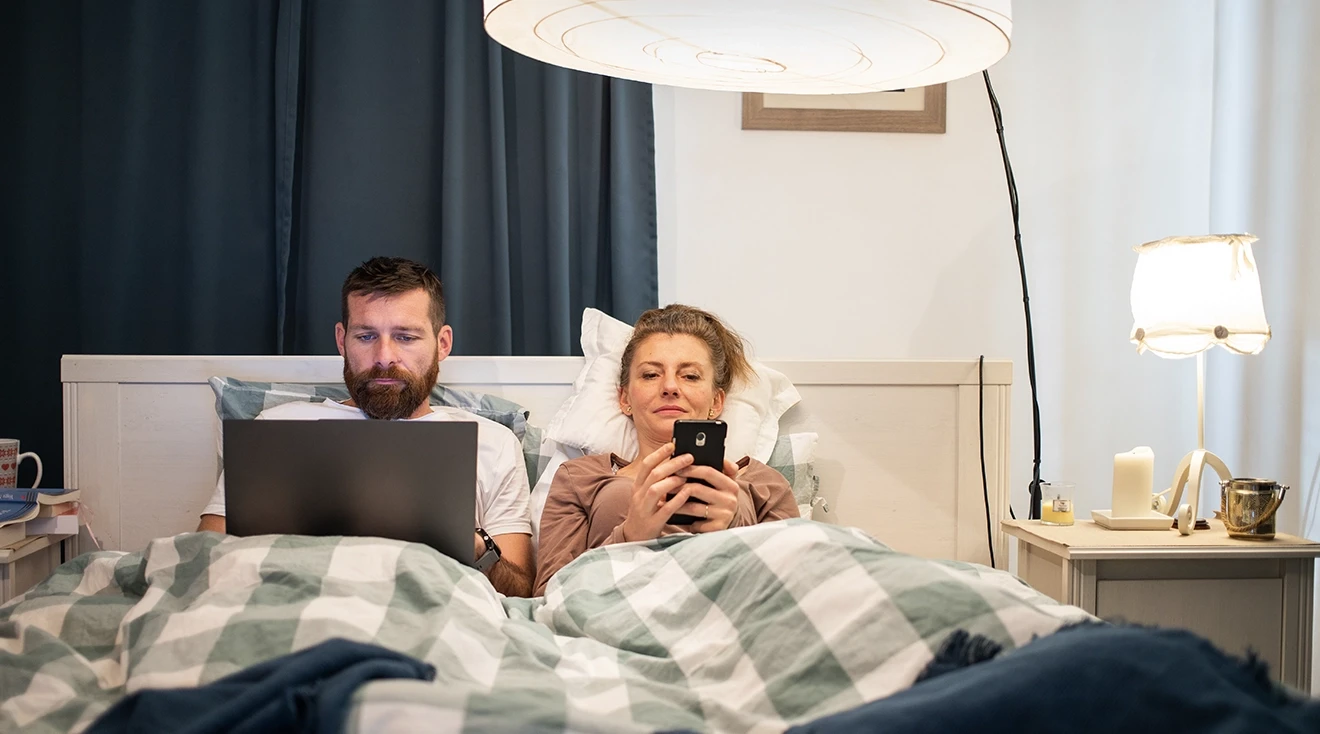 parents on laptop and phone while sitting in bed at home