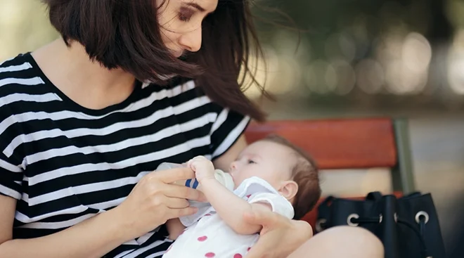 mother feeding baby a bottle on a bench in the park