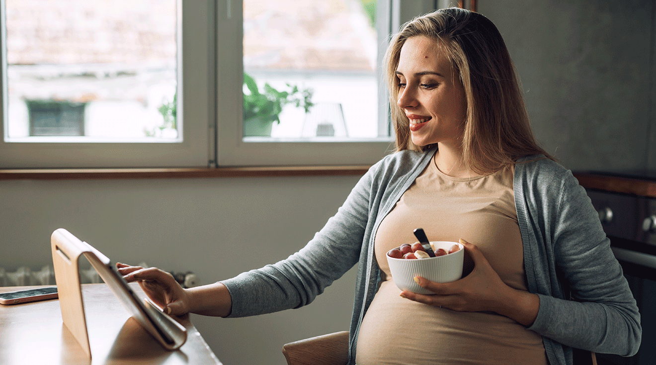 pregnant woman eating a snack while using tablet at kitchen table