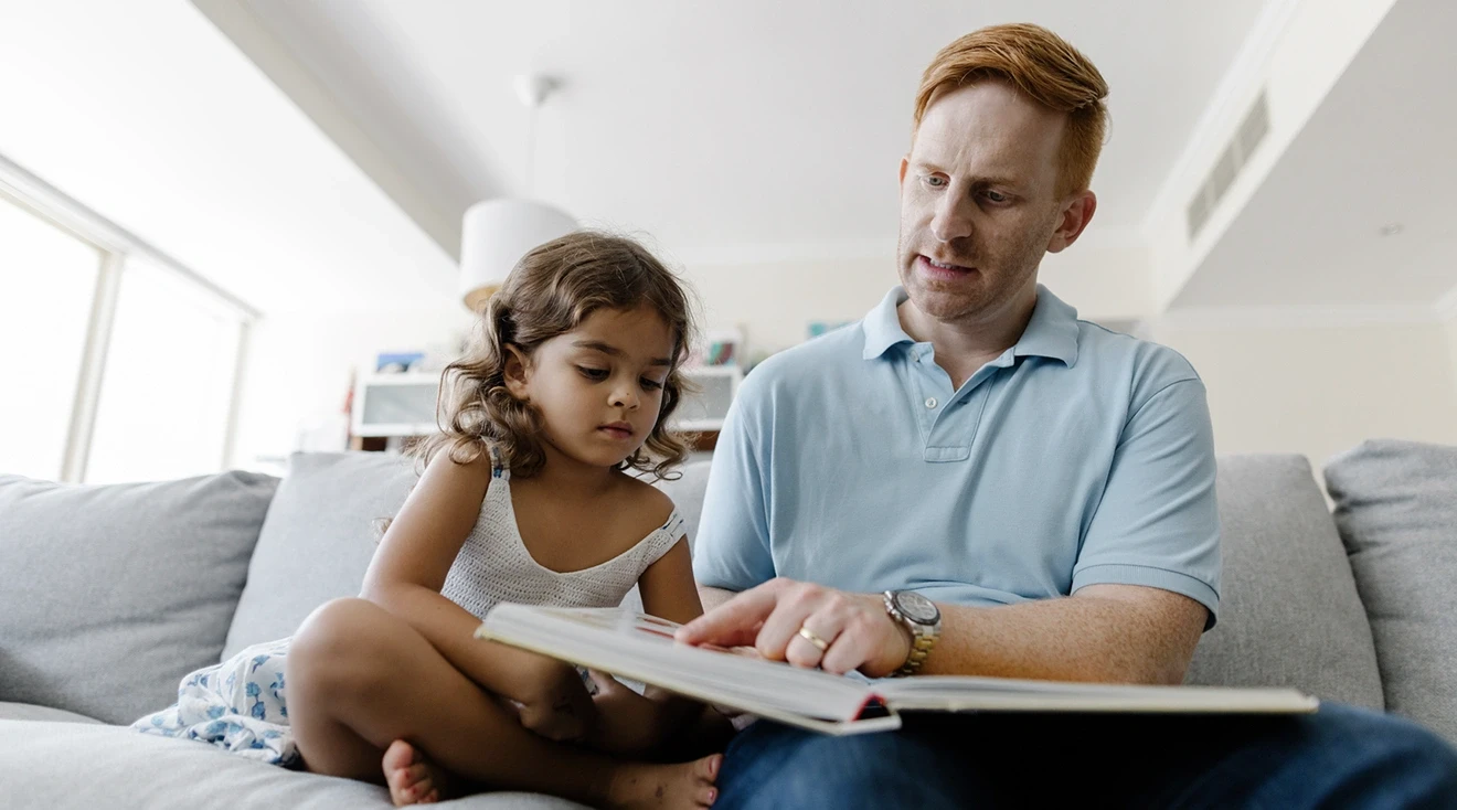 father looking at a book with young daughter while sitting on the couch at home