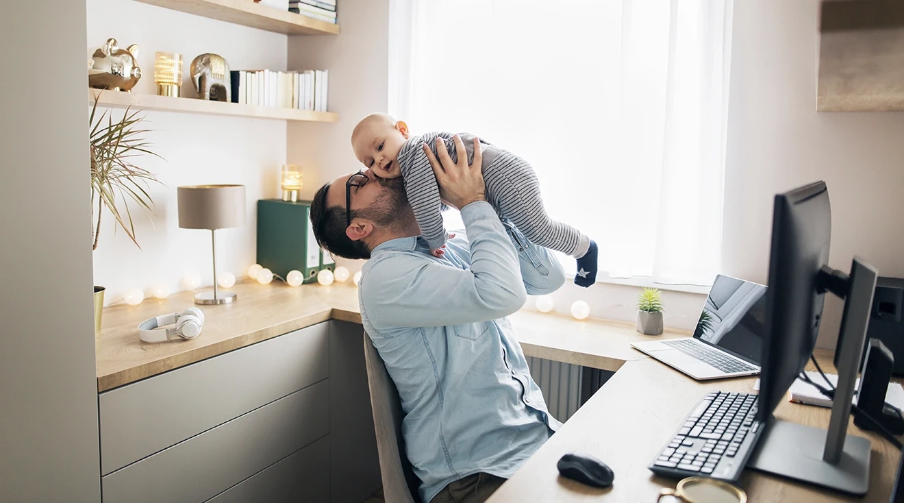 dad sitting at desk at home while holding baby