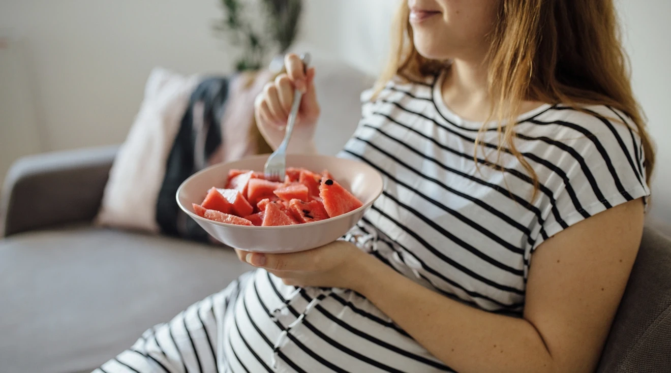 pregnant woman eating a bowl of watermelon on the couch at home