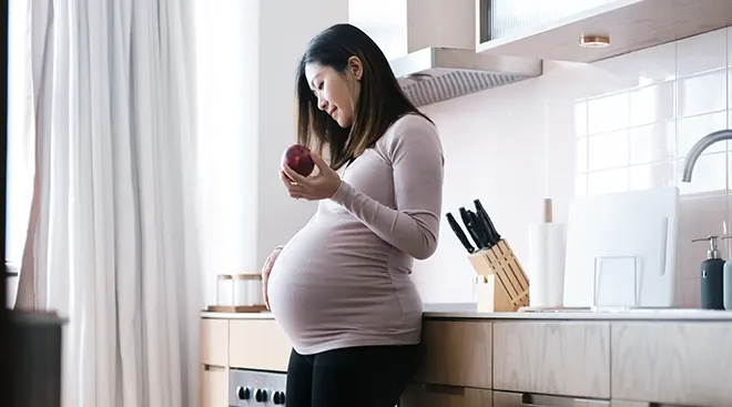 pregnant woman holding an apple while standing in kitchen at home