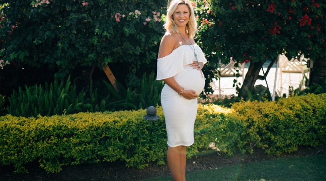 happy pregnant woman in white dress posing in front of flowers and shrubbery 