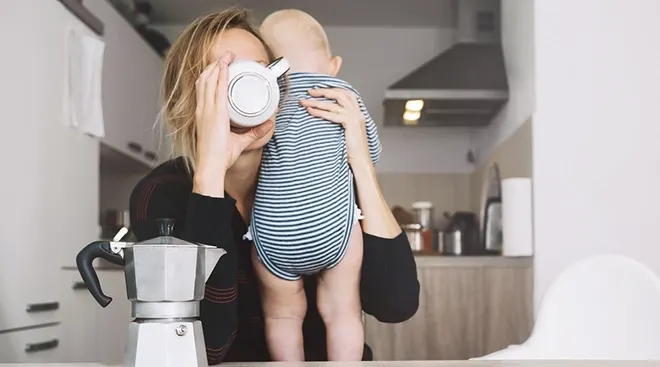 tired mother holding baby while drinking coffee at home