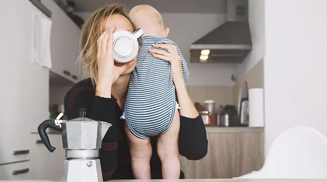 tired mother holding baby while drinking coffee at home