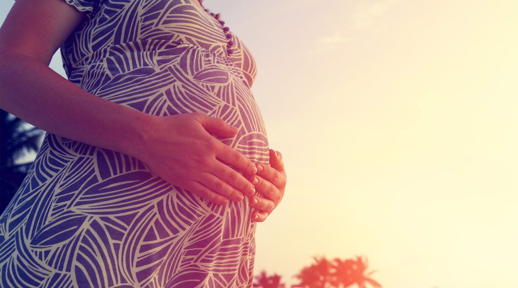 pregnant woman with hands on her belly, standing on the beach at sunset