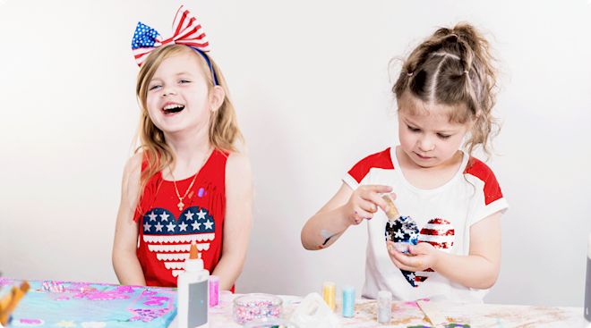 Two little girls wearing fourth of july outfits doing arts and crafts.