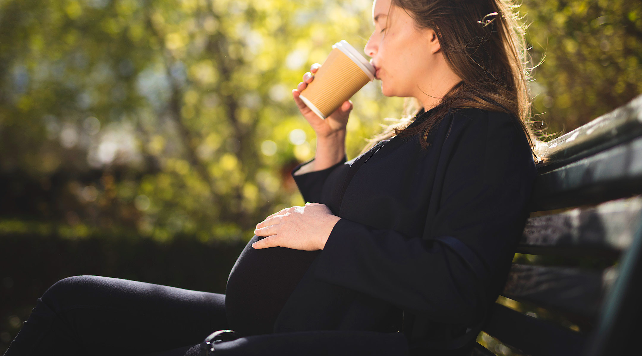 pregnant woman on a bench outside drinking coffee