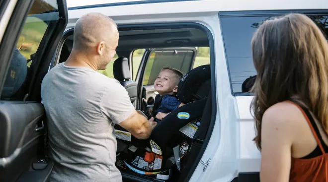 mother and father putting smiling toddler into car seat