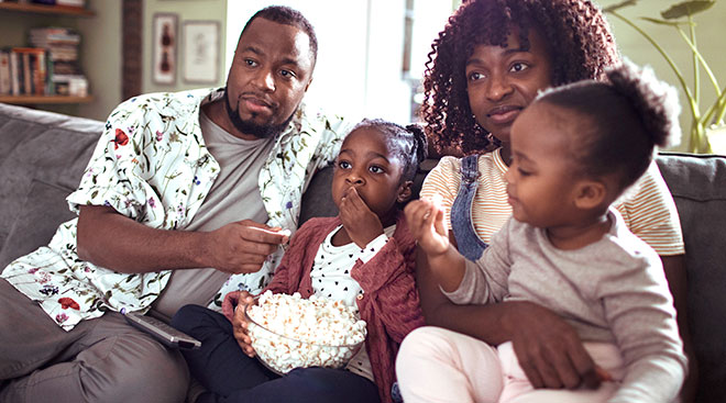 family sits on the couch together while eating popcorn and watching TV. 