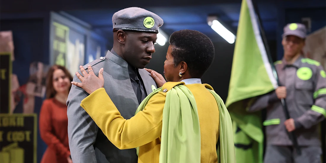 A person in a gray uniform and cap faces another person in a yellow jacket and cape, holding their shoulders, with others and green flags in the background.