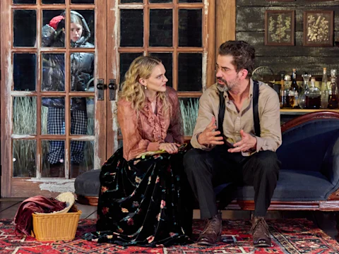 Production photo of The Disappear in New York, showing a woman and man sit talking on a sofa indoors, while a child in a coat looks in through glass doors from outside. Various bottles are on a side table behind them.