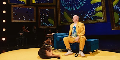 A person in a yellow suit sits on a blue couch on stage next to a dog, with musicians and colorful digital fireworks in the background.