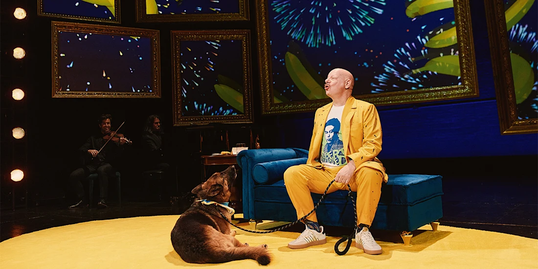 A person in a yellow suit sits on a blue couch on stage next to a dog, with musicians and colorful digital fireworks in the background.