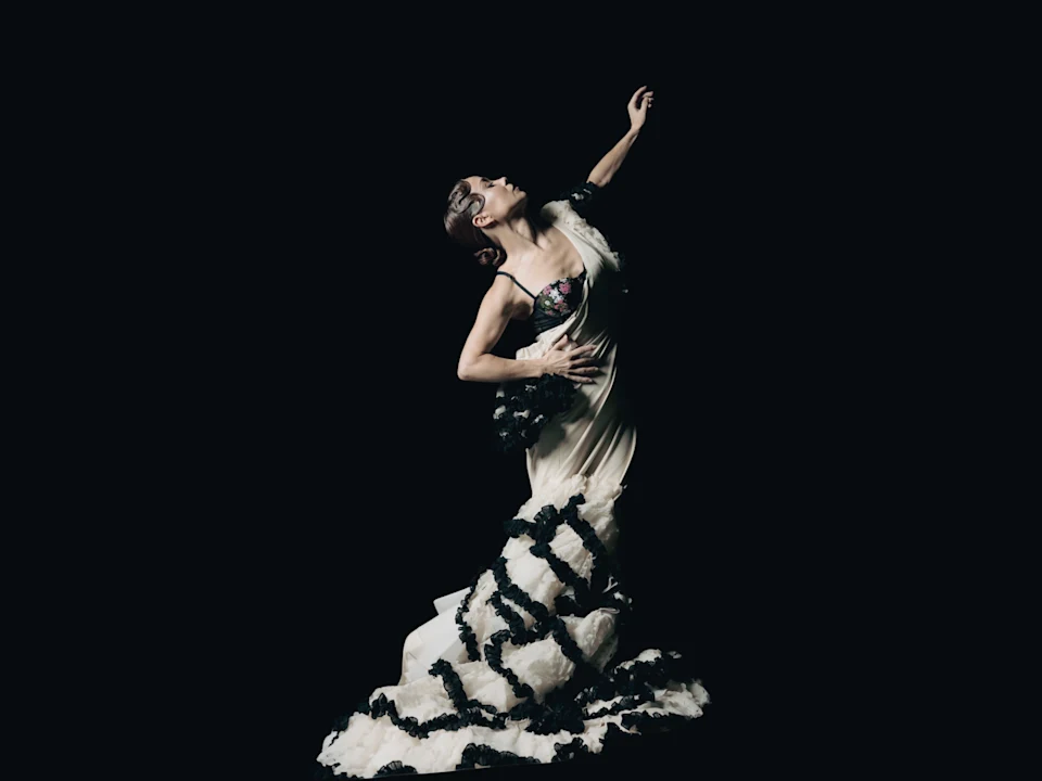 A person in a ruffled black and white dress poses dramatically against a black background, with one arm raised and head tilted back.