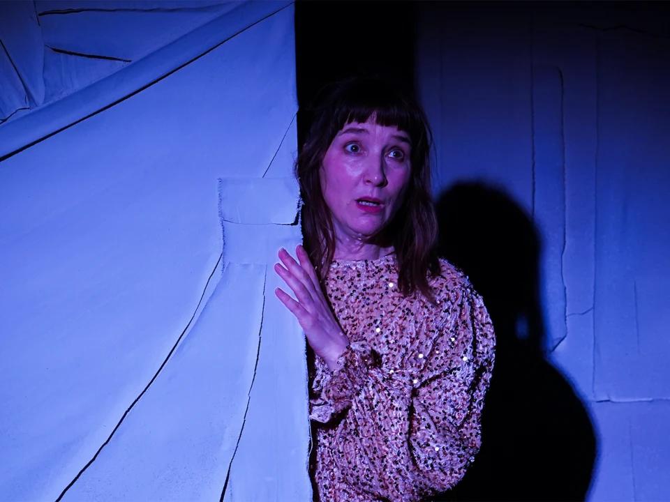 A person with long hair and a patterned shirt looks surprised as they peek out from behind a curtain in a dimly lit setting.
