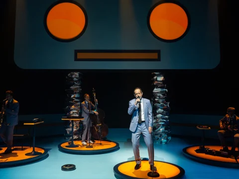 A band performs on stage with a futuristic robot face backdrop. The lead singer holds a microphone, and musicians play instruments on illuminated circular platforms.