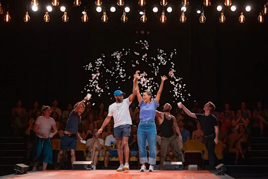 Five people on stage celebrate under confetti with their arms raised; an audience sits in the background, illuminated by overhead lights.