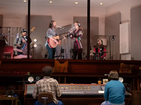 Five musicians perform in a recording studio, with two singing and playing guitar behind glass, while two people sit at a mixing console in the control room.