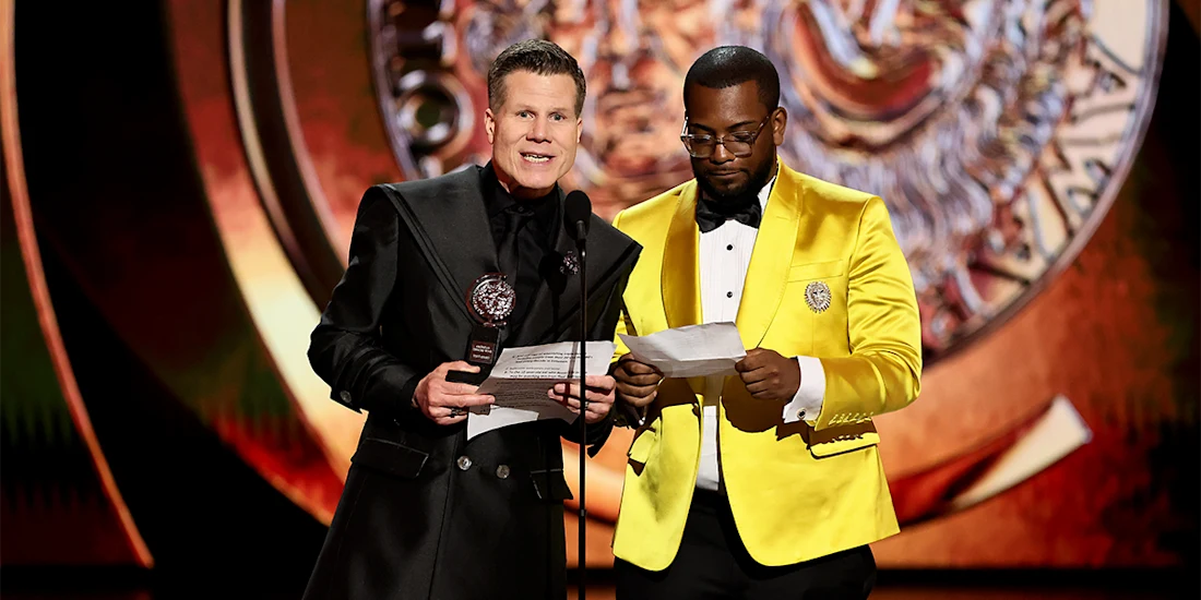 Two men stand on stage at an award ceremony, one holding a trophy and speaking, while the other reads from a sheet of paper.
