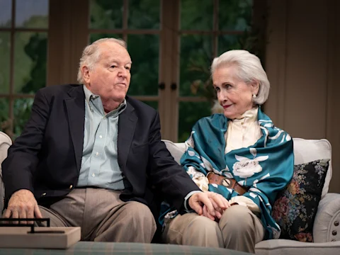 An older man and woman sit close together on a couch, holding hands and looking at each other, both dressed in formal attire.