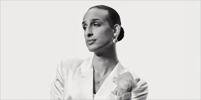 Person with slicked-back hair wearing a light-colored blazer and earrings, looking to the side against a plain background.