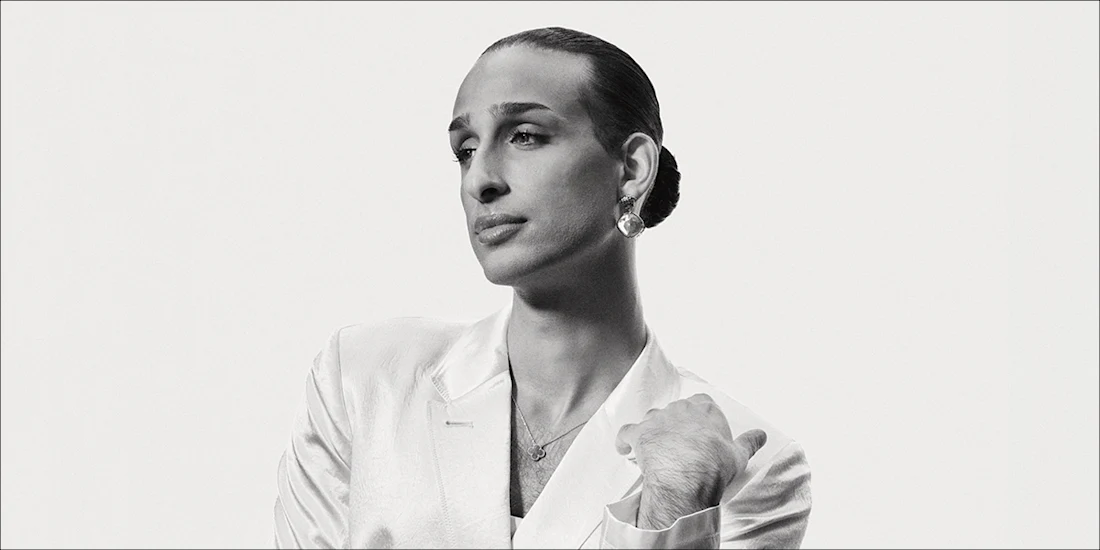 Person with slicked-back hair wearing a light-colored blazer and earrings, looking to the side against a plain background.