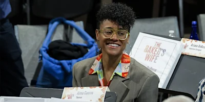 Person with short curly hair, large glasses, and a patterned shirt smiles while seated among signs for "The Baker's Wife" at an event.