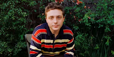 A person with short hair sits outdoors in front of green foliage, wearing a navy blue, red, and yellow striped sweater, and looks directly at the camera.