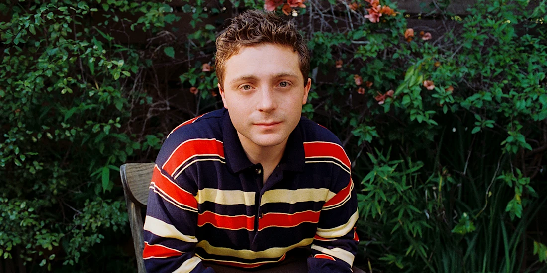 A person with short hair sits outdoors in front of green foliage, wearing a navy blue, red, and yellow striped sweater, and looks directly at the camera.