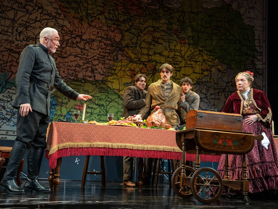 Five actors in period costumes perform around a table covered with food, set against a large map backdrop on stage.