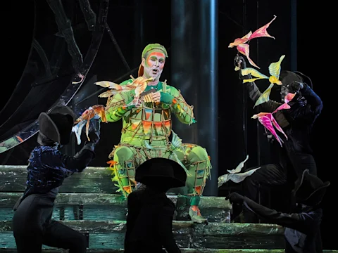 Production photo of The Magic Flute—Holiday Presentation in New York, showing a performer in a green costume sits on stage holding colorful bird props, surrounded by other actors dressed in dark outfits and hats during a theatrical production.