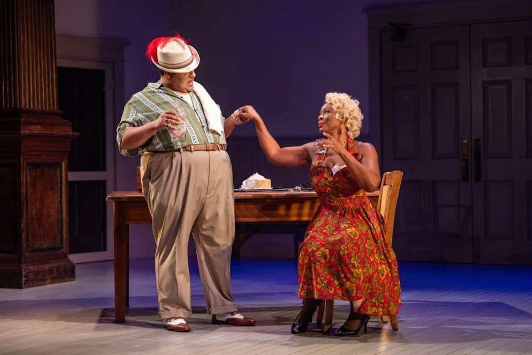 Two actors perform on stage; one stands in a hat and patterned shirt, offering a hand to a seated woman in a red floral dress beside a table with a dessert.
