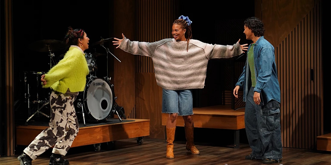 Three people stand on a stage in front of a drum set, smiling and interacting enthusiastically with each other.