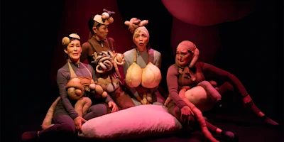 Four performers in flesh-toned costumes with exaggerated body part props sit on stage around a large pillow, under dramatic lighting.