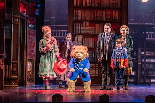 A group of adults and children stand on stage in a library setting with a person in a Paddington Bear costume at the center.
