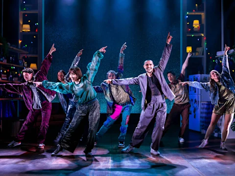 A group of performers on stage dance in coordinated poses, wearing colorful costumes under dramatic stage lighting, with set decorations and string lights in the background.