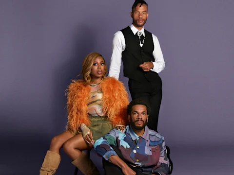 Three stylish people pose against a purple background; one stands in a vest and tie, the other two sit, one in an orange feathered jacket and boots, the other in a patterned shirt.