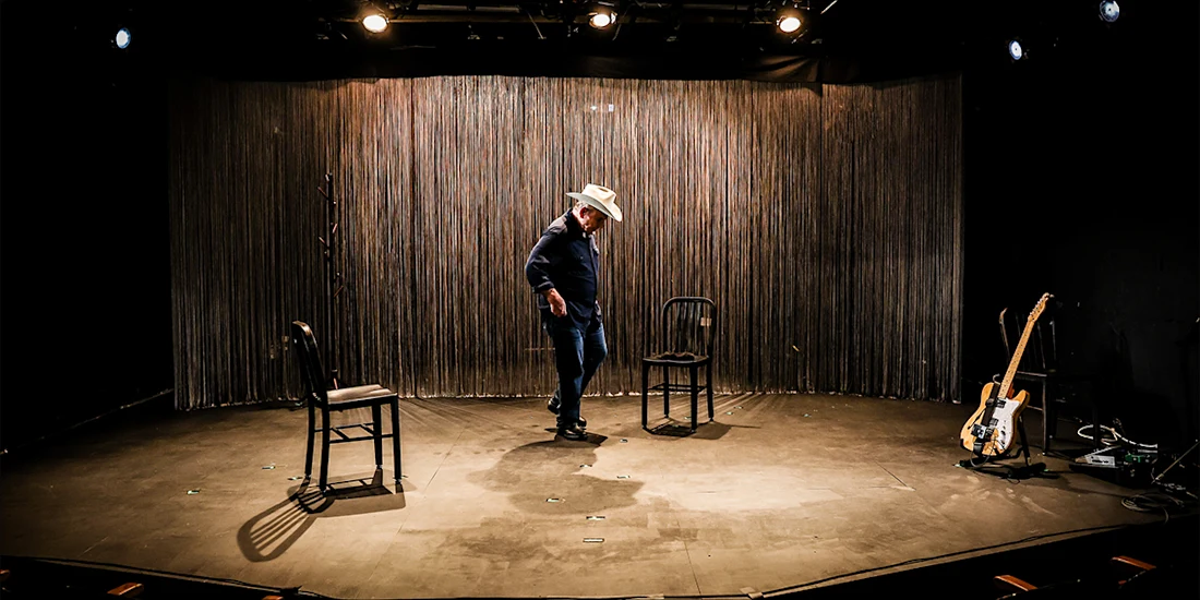 A man in a cowboy hat stands on a stage with three chairs and an electric guitar on the floor. Stage lights shine overhead.