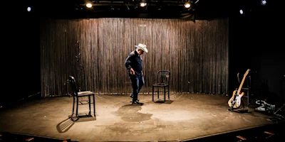 A man in a cowboy hat stands on a stage with three chairs and an electric guitar on the floor. Stage lights shine overhead.