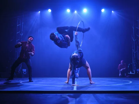 Performers execute an acrobatic routine under blue and purple lighting, with one person in mid-air above a hoop and another supporting from below. Two others observe from the sides of the stage.