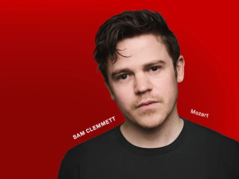 Photo of Sam Clemmett (Mozart) with short brown hair and slight stubble wearing a black shirt stands against a solid red background.