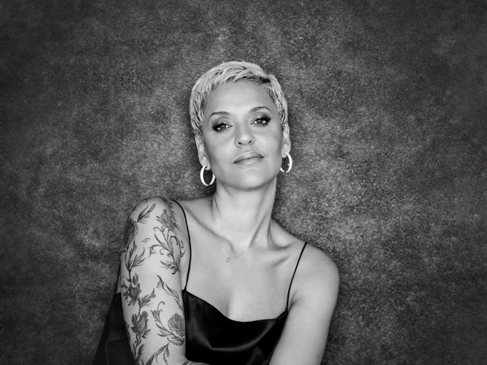 A woman with short hair and hoop earrings poses confidently against a textured backdrop. She wears a sleeveless top, revealing a detailed arm tattoo.