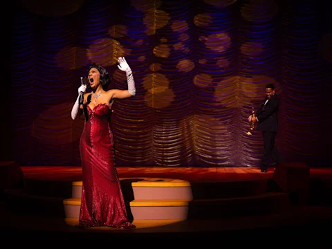 A woman in a red gown sings on stage with one arm raised, while a man in a suit holding a trumpet stands in the background. The stage is lit with dramatic lighting.