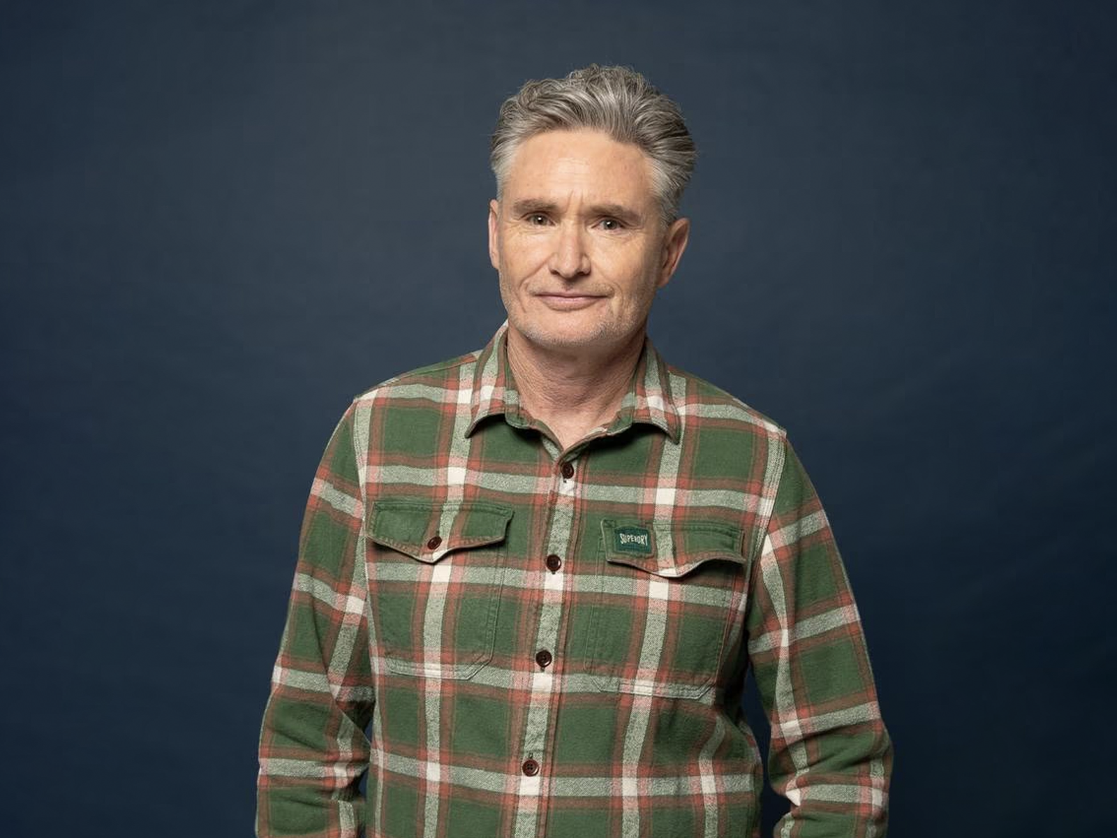 A middle-aged person with short gray hair stands in front of a dark blue background, wearing a green and red plaid button-up shirt.