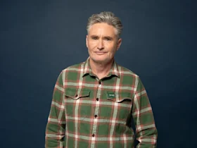 A middle-aged person with short gray hair stands in front of a dark blue background, wearing a green and red plaid button-up shirt.