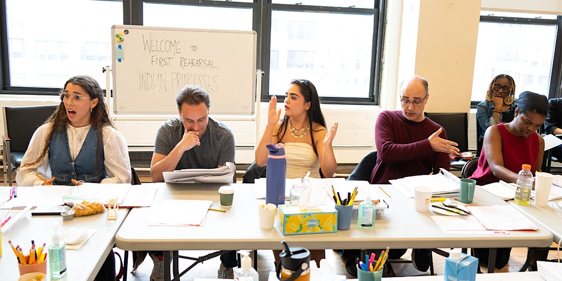 Five people sit at a table with scripts, drinks, and snacks, participating in a rehearsal; a whiteboard in the background reads, "Welcome to First Rehearsal Indian Princess.