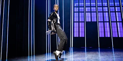 A performer in a black sequined jacket, white gloves, and black pants dances on stage with vertical blue lights and large windows in the background.