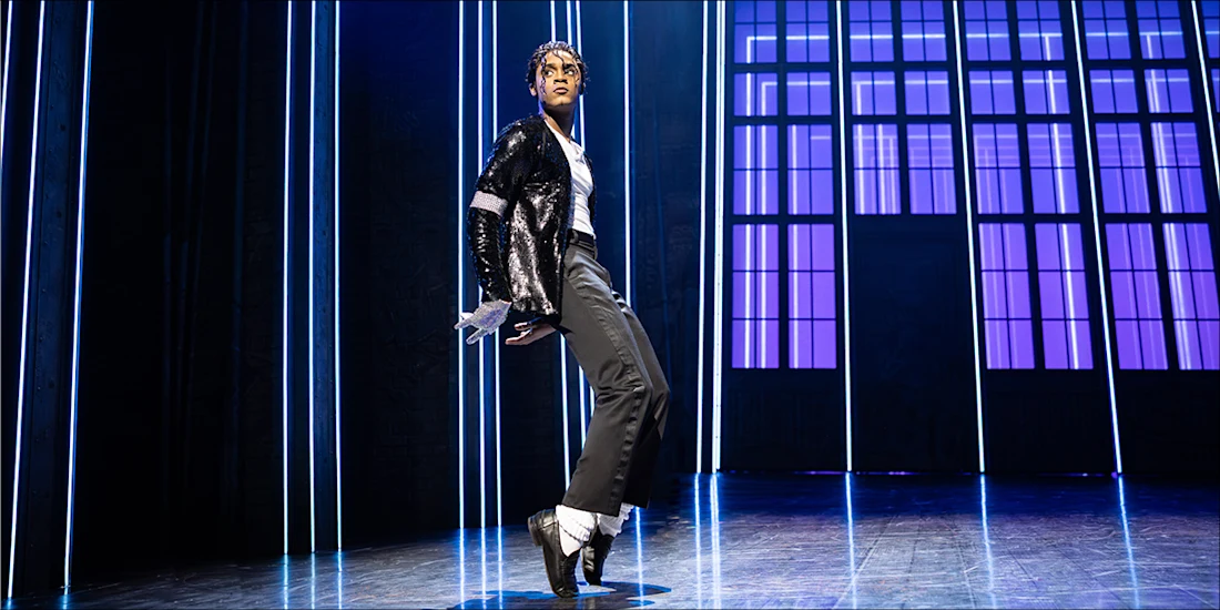 A performer in a black sequined jacket, white gloves, and black pants dances on stage with vertical blue lights and large windows in the background.